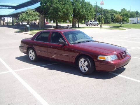 2004 Ford Crown Victoria - Dark red Ford Crown Victoria sedan photographed from the passenger-side front three-quarter angle in an empty parking lot, wearing a blacke…