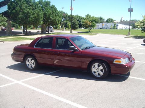 2004 Ford Crown Victoria - Passenger-side three-quarter front view of a dark red Ford Crown Victoria sedan parked across the stalls of an empty asphalt lot on a sunny…