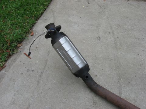 1994 Ford Bronco - A removed exhaust catalytic converter sits on the ground, showing its metallic casing and connecting pipes.
