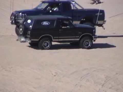 1995 Ford Bronco - A black Ford Bronco is parked in front of a dark blue Ford pickup truck on a sandy surface.