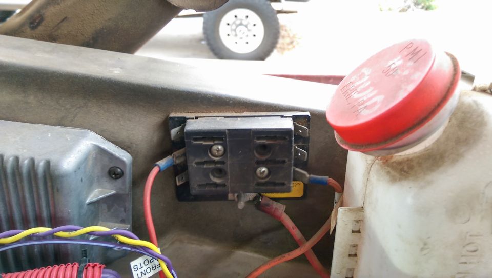 Close-up of a vehicle's fuse box with red and blue wires connected, next to a red fluid reservoir cap with text