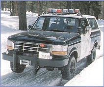 1995 Ford Bronco - Small photo of a black-and-white full-size Ford Bronco in police-style livery with a roof light bar, spotlight, and winch bumper, driving a…