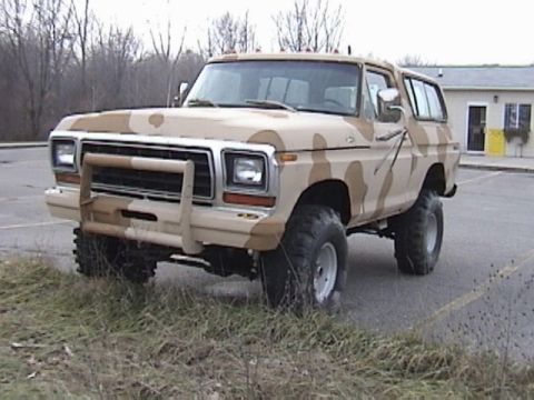 1995 Ford Bronco - Driver-side three-quarter front view of a late-1970s full-size Ford Bronco painted in tan and brown desert camouflage, parked at the edge o…