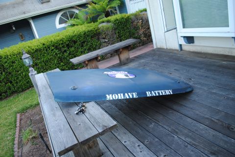 1986 Ford Bronco - A blue car hood with the text "MOHAVE BATTERY" and a circular emblem is resting on a wooden deck next to a hedge and a house.