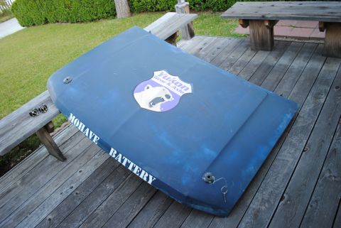 1986 Ford Bronco - A dark blue vehicle hood is laid out on a wooden deck, featuring a 'Yukon Gear & Axle' sticker and 'MOHAVE BATTERY' text in white. A small…