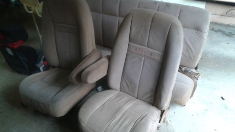 1996 Ford Bronco - Two tan fabric bucket seats with armrests and a matching bench seat are displayed in a garage setting. The bucket seats have a decorative s…