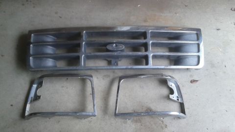 1996 Ford Bronco - A chrome grille and two chrome headlight bezels are laid out on a concrete surface. The grille has horizontal bars and a central oval emble…