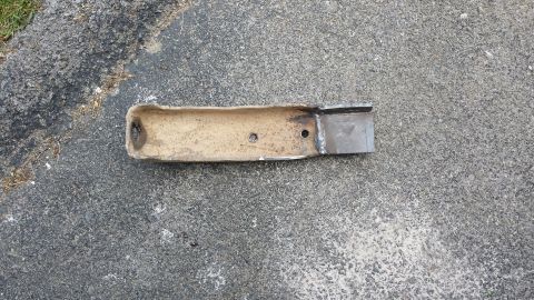 Rusted hollow metal exhaust pipe section with holes and a welded plate on asphalt.