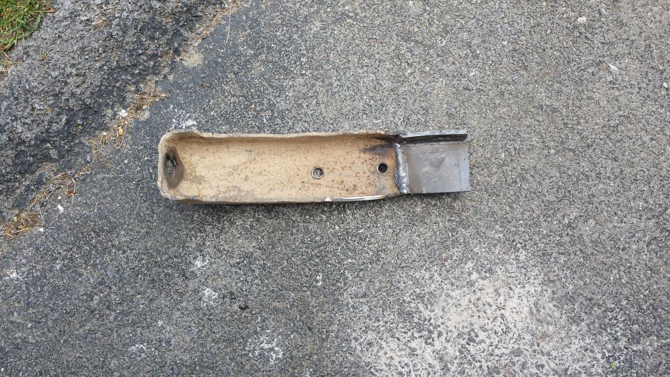 Rusted hollow metal exhaust pipe section with holes and a welded plate on asphalt.
