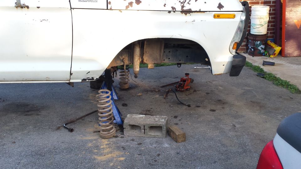 White 1970s Ford truck/SUV front passenger side, suspension disassembled, coil spring removed, jack and jack stands visible.