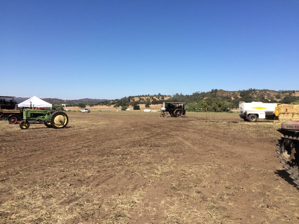 Green vintage tractor in a field with other vehicles and hay bales.