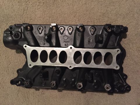 1996 Ford Bronco - A black cast iron intake manifold with a silver gasket is shown from a top-down perspective, resting on a beige carpeted surface. Visible t…