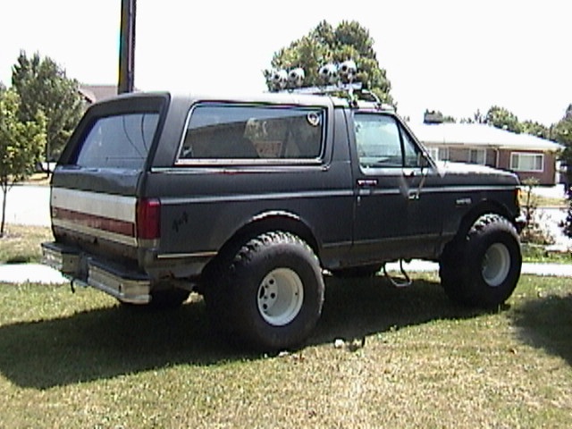 Lifted black Ford Bronco with white/red stripe, big tires on white wheels, and roof lights, parked on grass