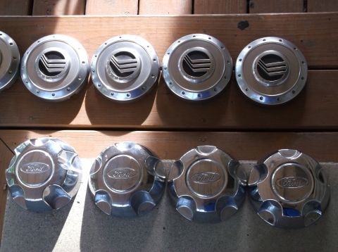 1999 Mercury Grand Marquis - Two rows of four car hubcaps are displayed on a wooden deck. The top row features four metallic hubcaps with a circular design and a V-shap…