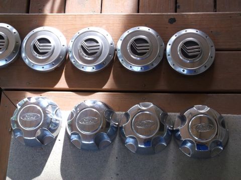 1999 Mercury Grand Marquis - Four chrome Ford branded hubcaps are shown in the foreground, with four silver Mercury branded hubcaps arranged above them on a wooden surf…