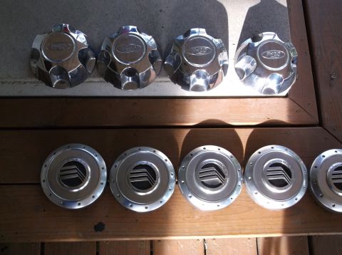 1999 Mercury Grand Marquis - Four chrome Ford hubcaps are displayed on a wooden surface in the top row, and six silver Mercury hubcaps are arranged below them.