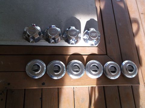 1999 Mercury Grand Marquis - A collection of seven chrome-finished wheel covers are displayed, with four on a wooden surface and three on a concrete surface.