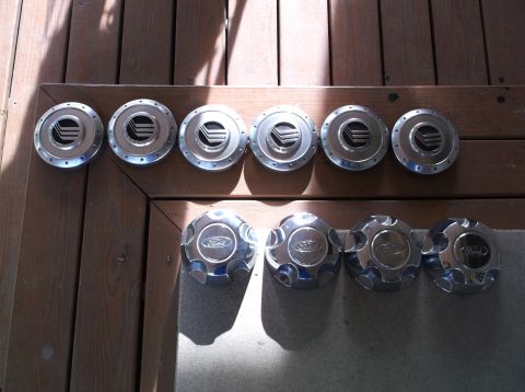 1999 Mercury Grand Marquis - Two rows of car hubcaps are displayed on a wooden surface. The top row features five silver hubcaps with a black emblem, and the bottom row…