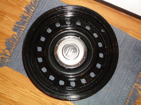 1999 Mercury Grand Marquis - A black Mercury hubcap with a silver center cap featuring the Mercury logo is shown on a wooden floor with a blue rug. The hubcap has a pat…