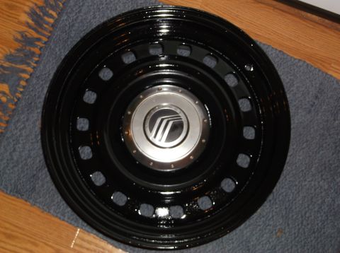 1999 Mercury Grand Marquis - A black hubcap with a silver center cap featuring the Mercury logo is displayed on a dark rug. The hubcap has multiple square cutouts aroun…