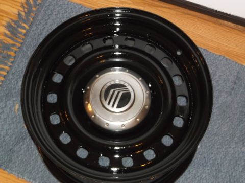 1999 Mercury Grand Marquis - A black steel wheel with a silver Mercury logo center cap is shown, resting on a blue rug and wooden floor.