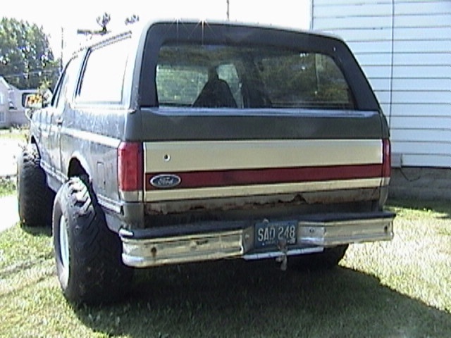 Gray Ford Bronco rear view on grass, rusty tailgate, big mud tires, roof light bar