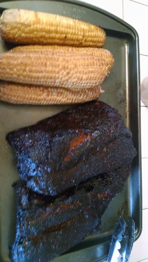 1979 Ford Bronco - Three ears of corn and two racks of ribs are arranged on a dark baking sheet. Tongs are visible on the right side of the baking sheet.