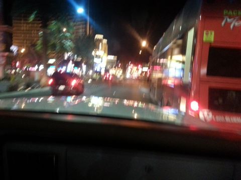 1979 Ford Bronco - A red car is seen from inside another vehicle driving through a brightly lit city at night.