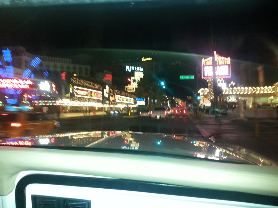 Dashboard view from inside a vehicle looking out at a brightly lit street at night with neon signs and traffic