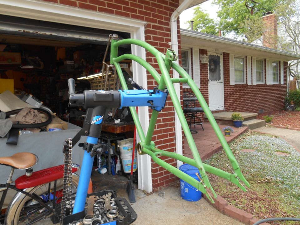 Green bicycle frame clamped in a blue Park Tool repair stand, with a garage and house in the background
