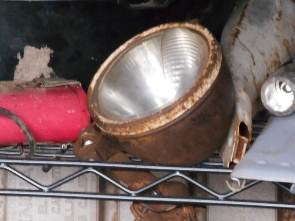Rusty headlight assembly with ribbed glass lens, next to a red cylindrical object