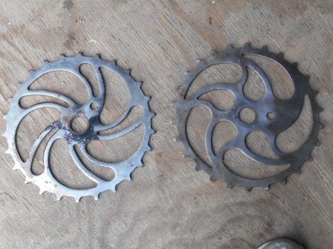 Two metal bicycle chainrings on a wooden surface. The left chainring is silver with black wear marks. The righ