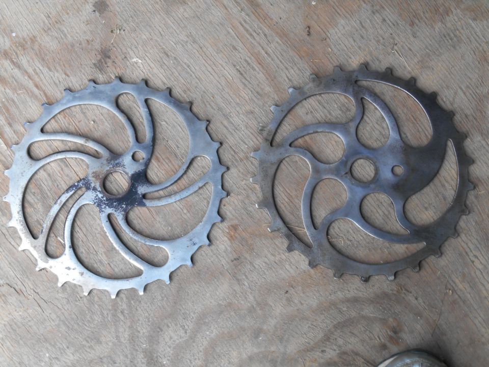 Two metal bicycle chainrings on a wooden surface. The left chainring is silver with black wear marks. The right chainring is darker gray
