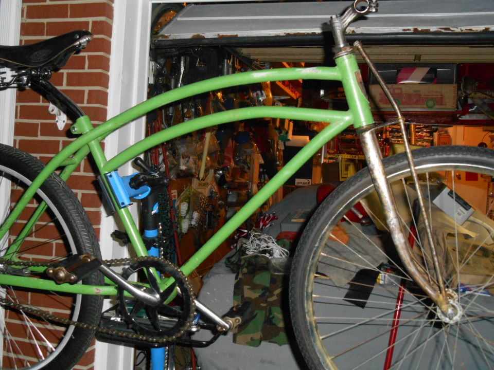 Green cruiser bicycle frame with a black seat and chrome handlebars. The bike is in a garage with tools and supplies in the background