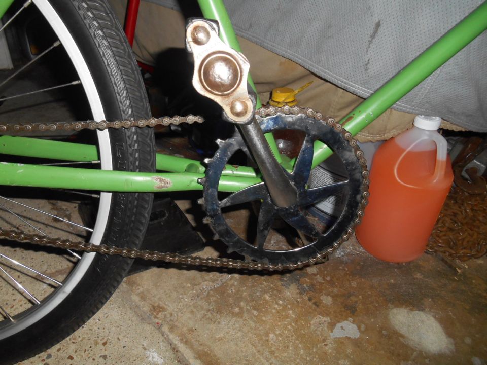 Green bicycle frame with chain, crankset, and rear wheel visible. Orange liquid in a jug sits nearby