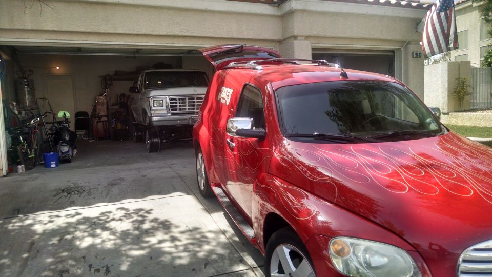 Silver pickup truck with a large grille parked in a garage. A red car with flame decals is in the foreground