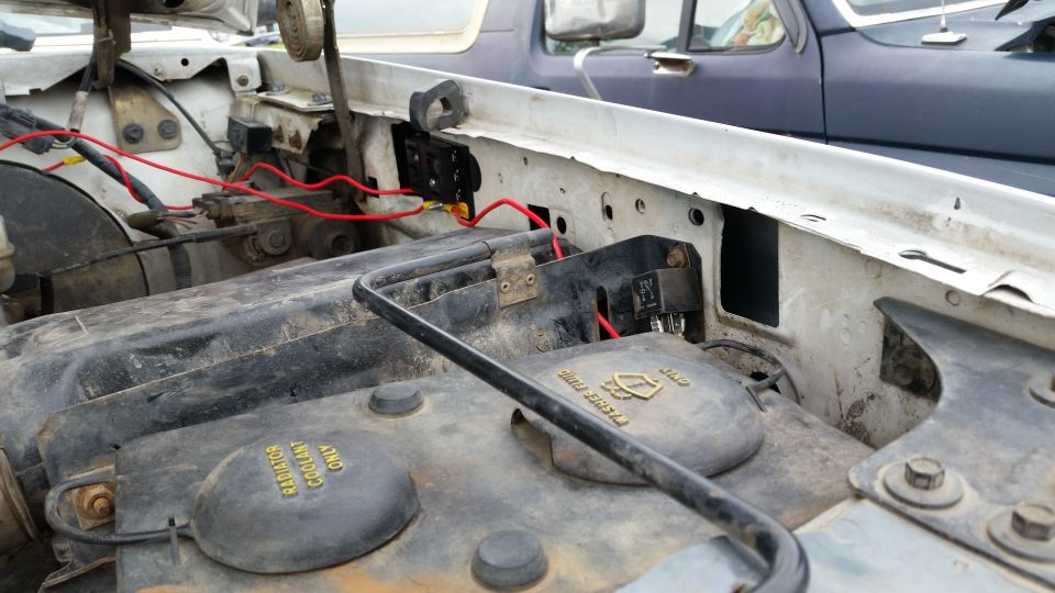Engine bay with red wires and coolant caps. Another vehicle is visible in the background