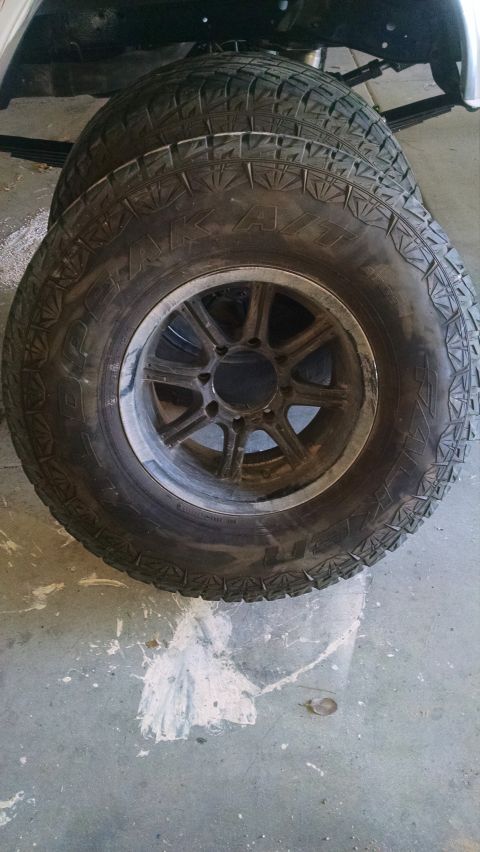 1979 Ford Bronco - A stack of four off-road tires with aggressive tread patterns are shown mounted on dark-colored rims. The tires are resting on a concrete f…