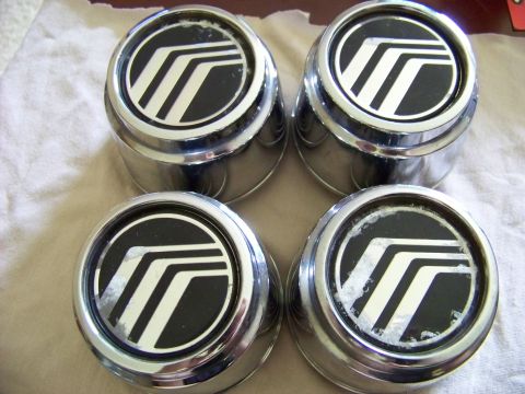 1999 Mercury Grand Marquis - Four chrome hubcaps with the Mercury logo are displayed on a light-colored surface. The hubcaps show some wear and tear on the black emblem.