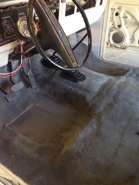 1978 Ford Bronco - The interior of a vehicle shows a dark grey carpet, a steering wheel with a Ford logo, and pedals. The driver's side door panel is visible…