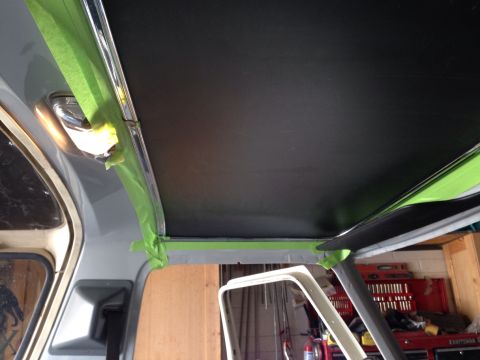 1978 Ford Bronco - The interior of a vehicle shows a black headliner being installed, with green painter's tape protecting the surrounding trim and gray paint…