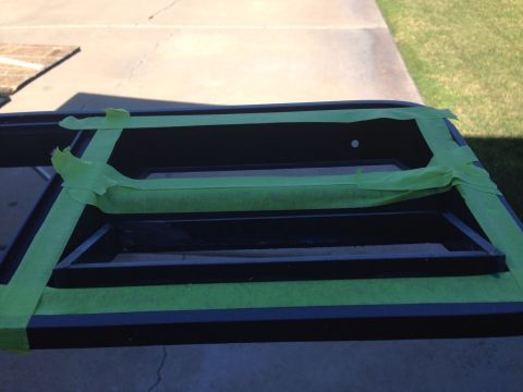 1978 Ford Bronco - A black vehicle part, possibly a grille or bumper insert, is covered with green painter's tape. The part is outdoors on a paved surface.