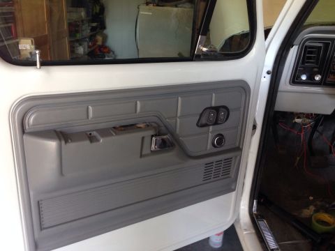 1978 Ford Bronco - The interior of a white Ford truck door is shown, with a gray door panel featuring a chrome door handle, window controls, and speaker grill…