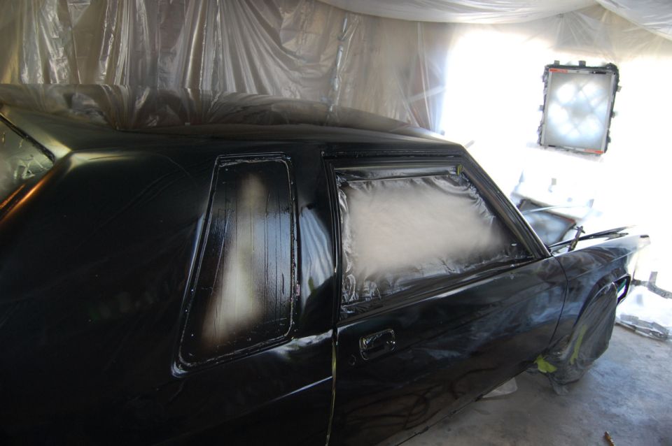 Black sedan being prepped for painting in a spray booth, windows taped off.
