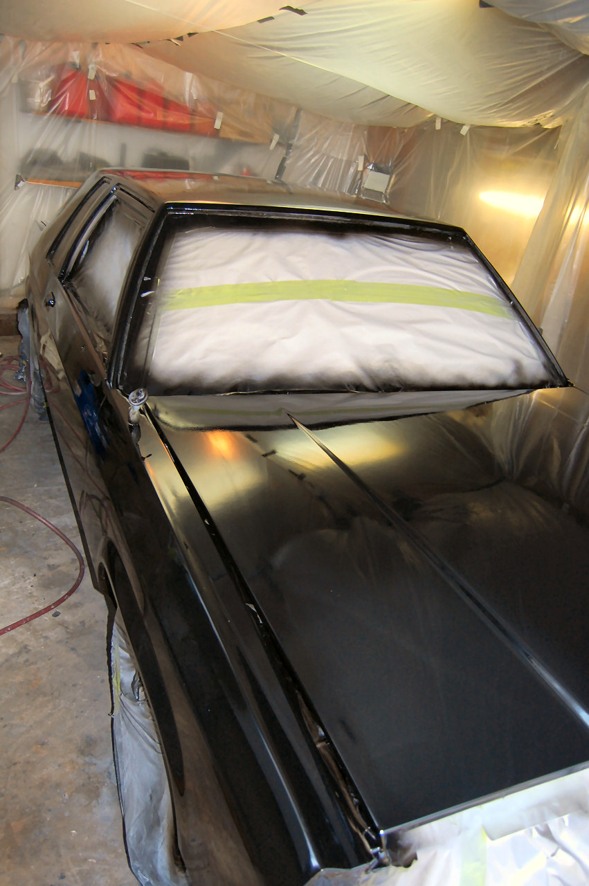 Black car bumper being painted, covered in plastic with green tape.