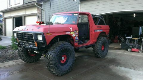 1994 Ford Bronco - A heavily modified red Ford Bronco with oversized tires and a roll cage sits parked in a driveway in front of a garage. The vehicle has num…