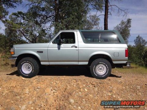 1994 Ford Bronco - The side view of a silver 1994 Ford Bronco with a green roof is parked on a dirt road with trees in the background.