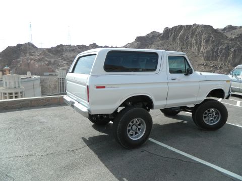 1979 Ford Bronco - A lifted, white 1979 Ford Bronco is parked on asphalt with a dam and rocky mountains in the background. The vehicle is viewed from the rear…