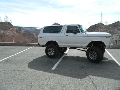 1979 Ford Bronco - A lifted white 1979 Ford Bronco is parked in a paved lot with a view of a large bridge and dam in the background.