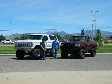 1979 Ford Bronco - Two men stand next to a lifted white Ford Bronco and a lifted maroon Ford F-150 pickup truck parked on a paved road with mountains in the b…